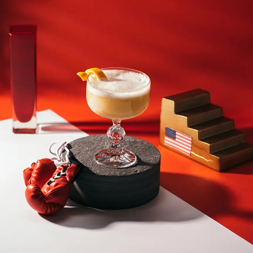 A glass of Nightcap Champions, a Cointreau triple sec cocktail, on a black platform with a pair of boxing gloves
